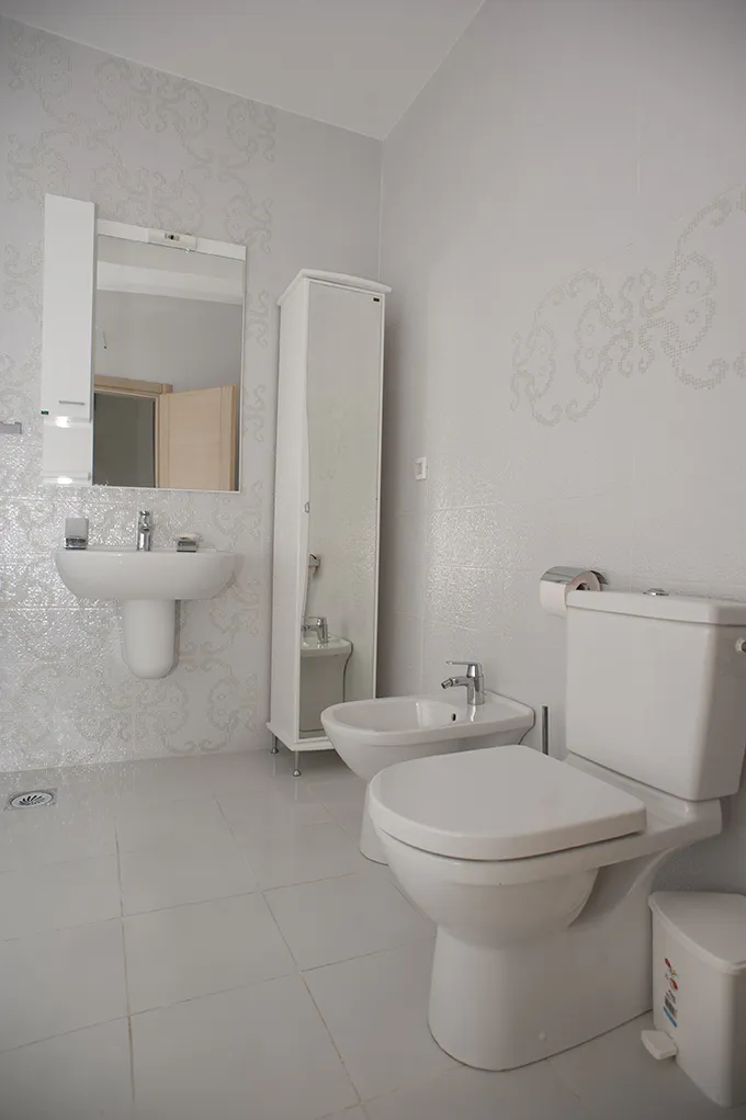 Bathroom featuring a toilet, bidet, wall sink, mirror, and white tiled walls with patterns.