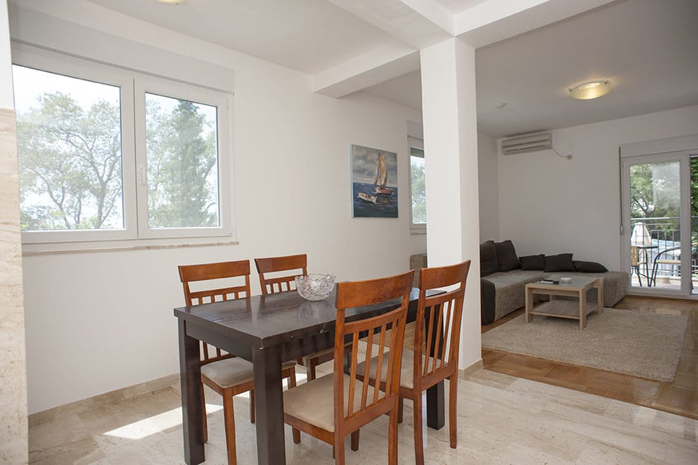 Living room with dining area in a villa in Perazica Do, Montenegro: dark wooden table, light furniture, and a view of the garden.