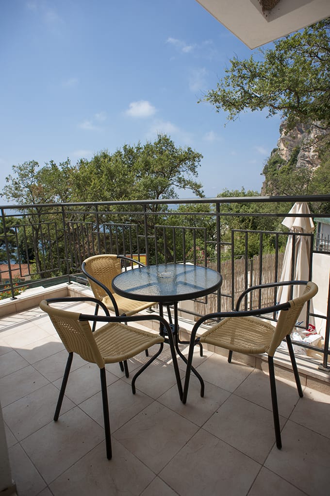 Villa balcony in Perazica Do, Montenegro: wicker chairs, a glass table, and a panoramic view of the sea and cliffs.