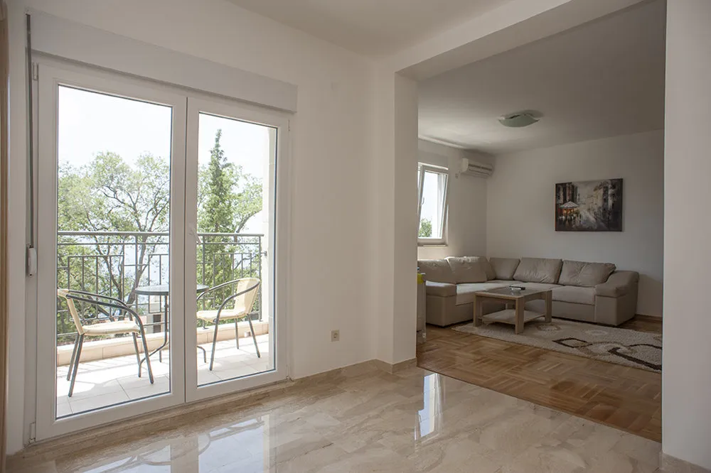 Living room with beige sofa and wooden table on parquet floor, viewed from a tiled hallway near balcony doors.