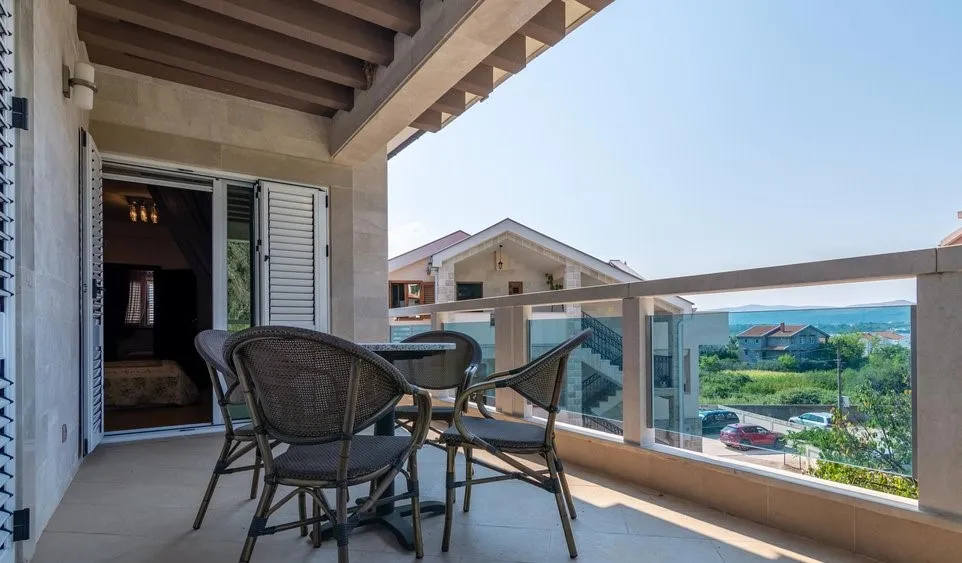 Open balcony with wicker furniture and glass railing, overlooking neighboring houses and hills.
