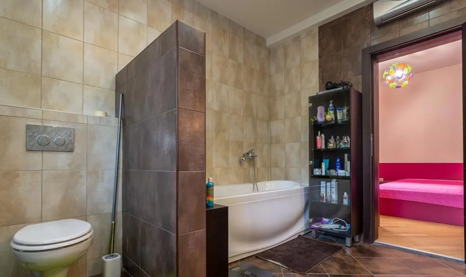 Bathroom featuring a white bathtub, toilet, and shelving with toiletries against beige tiled walls.