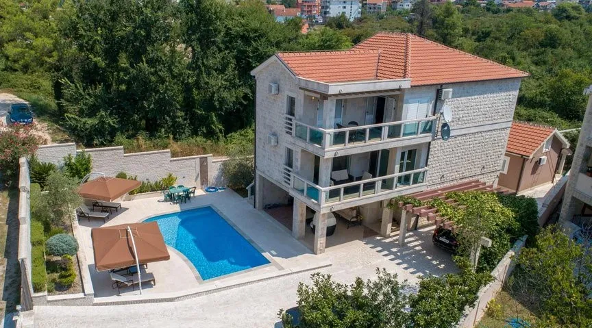 Aerial view of a house with a blue swimming pool, sun loungers under umbrellas, and surrounding greenery.