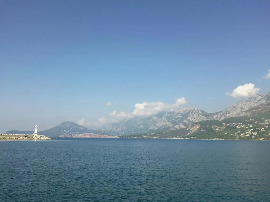 Seascape featuring a white lighthouse on a stone breakwater with a rugged mountain range under a clear blue sky.