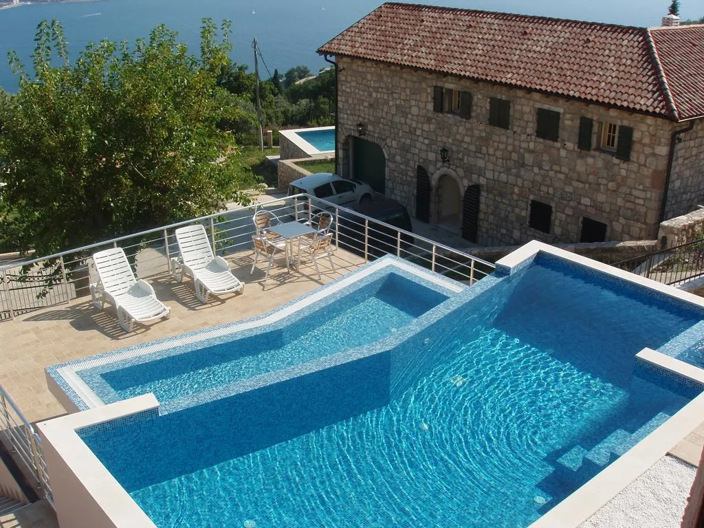 Blue tiled swimming pool on a terrace next to a stone house with sea view and white loungers.