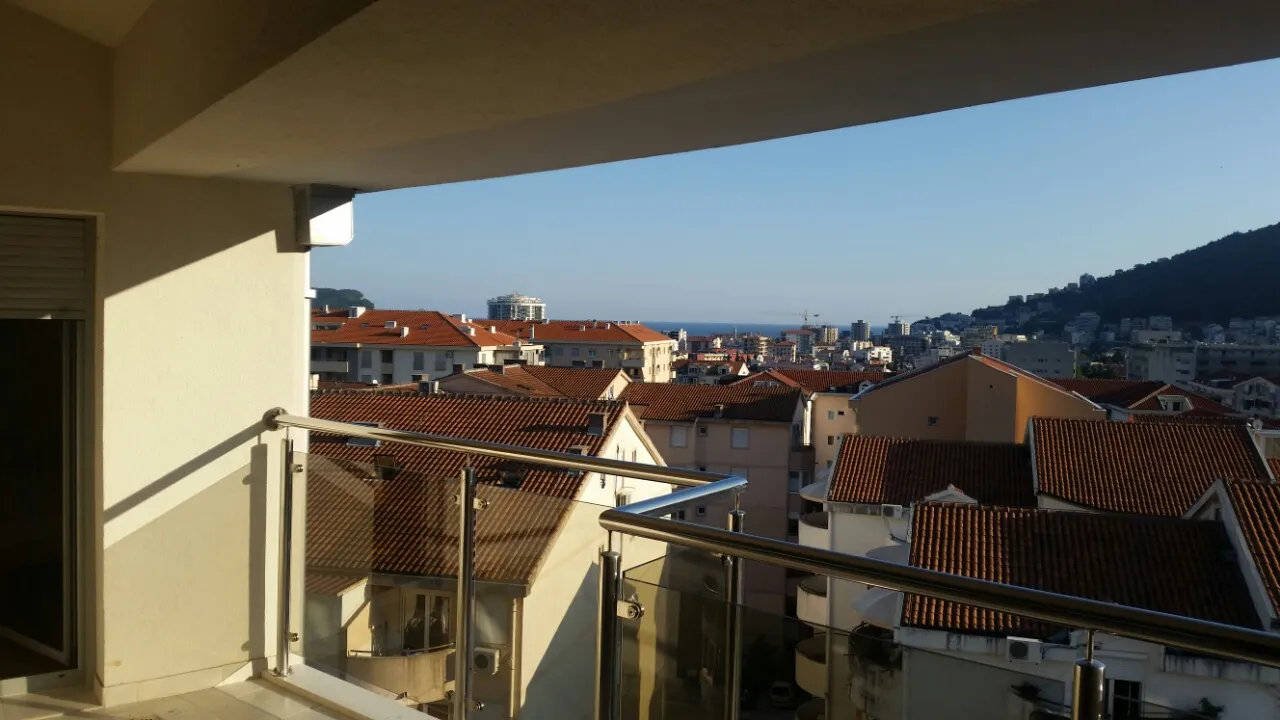 View from a balcony overlooking city rooftops and the sea. Features glass railings, a wall, and a door frame.