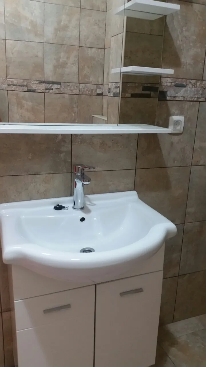 White sink with cabinet and chrome faucet against beige tiled wall featuring a mirror unit.