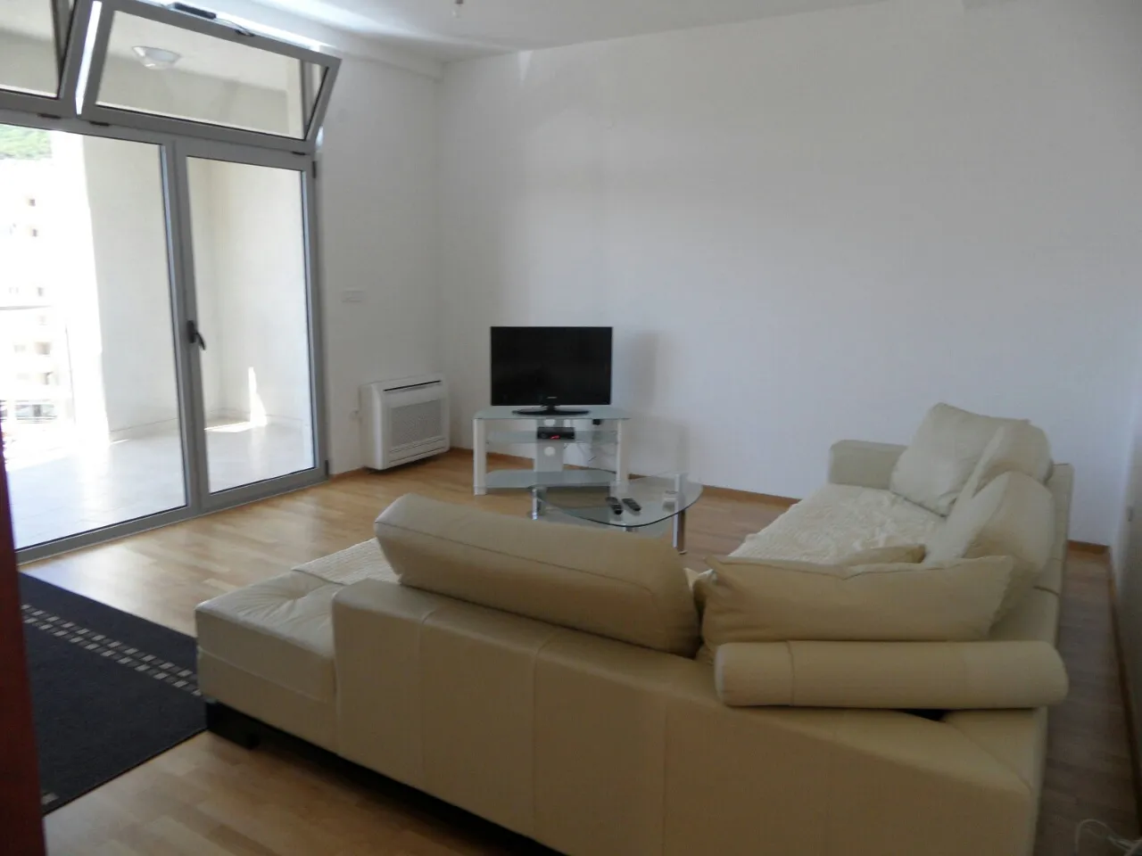Bright room with a beige sofa, TV on a glass stand, and large glass doors leading to a balcony.