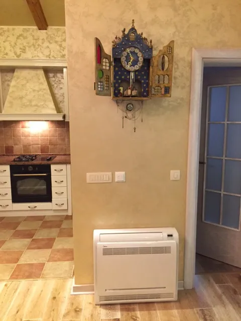 Partial view of a kitchen with stove, oven, and white cabinets. Decorative clock on wall, convector heater on floor.