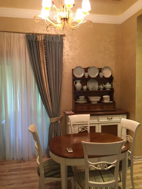 Dining area with a round table, white chairs, and a wooden hutch displaying dishes near curtains.