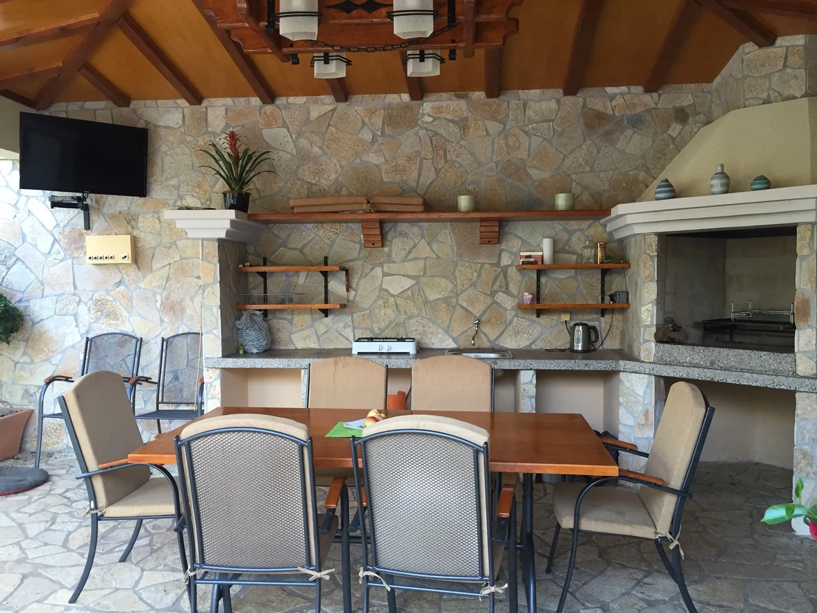 Open terrace with stone wall, wooden table, chairs, and built-in kitchen area featuring a barbecue grill.