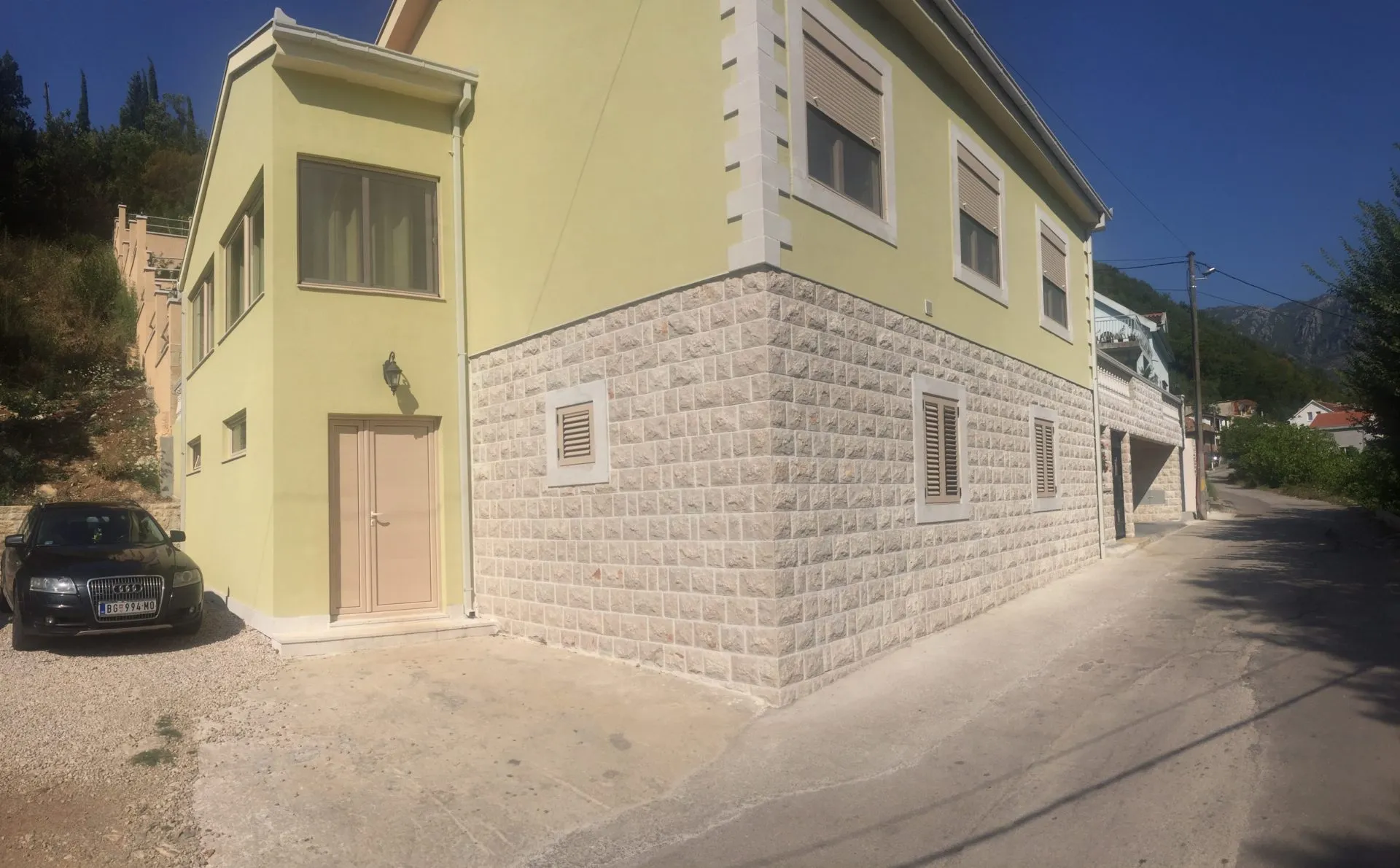 Exterior view of a two-story house with yellow walls and stone base. A black car is parked next to the entrance.