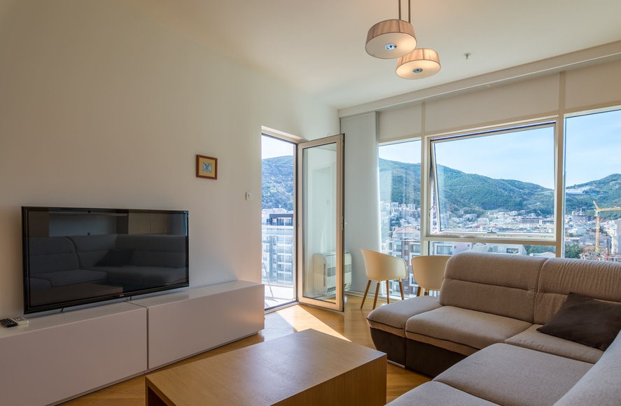 Modern living room in an apartment in Budva, Montenegro: panoramic windows with mountain views, light-colored furniture, and a TV area.