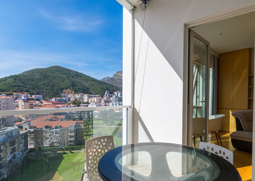 Apartment balcony in Budva, Montenegro: glass table, view of the mountains and the city, panoramic glazing, and access to the living room.