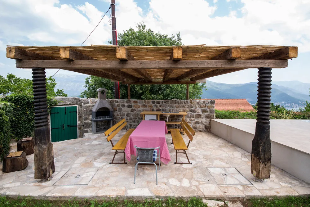 Open stone terrace with a wooden pergola, dining table in pink cloth, and outdoor oven against a mountain backdrop.