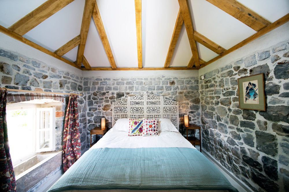 Bedroom in a stone villa on the Luštica Peninsula, Montenegro: a bed with a carved headboard and wooden beams on the ceiling.