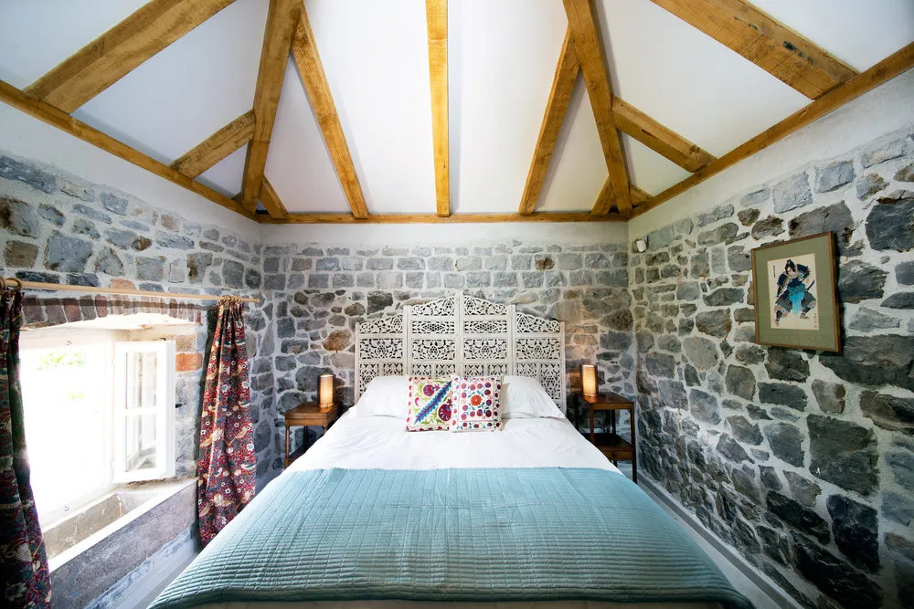 Bedroom featuring stone walls, exposed wooden ceiling beams, and a double bed with an ornate white headboard.