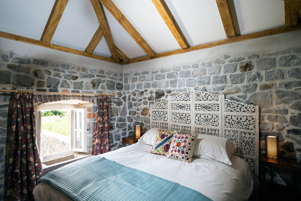 Bedroom in a villa in Luštica, Montenegro: stone walls, wooden beams, carved headboard, and a view from the window.