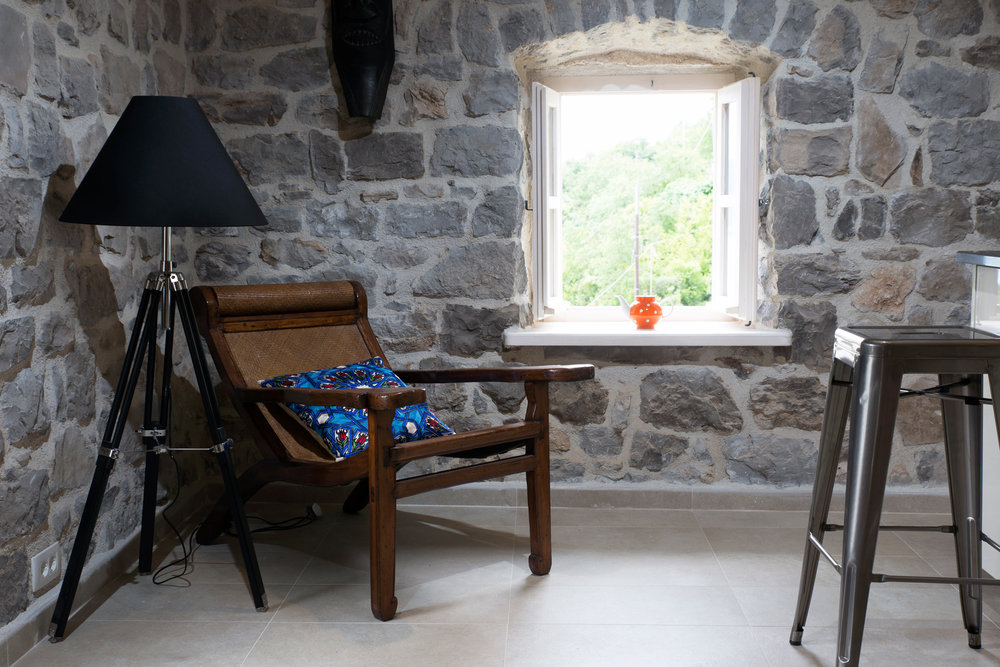 Living room interior in a villa in Luštica, Montenegro: stone walls, a vintage armchair, a floor lamp, and a view of the garden from the window.