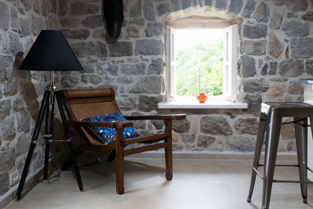 Room with stone wall, wooden armchair, black tripod lamp, and metal stool near an open window.