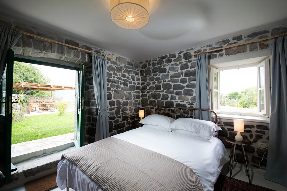 Bedroom with stone walls, double bed, nightstands, and open door leading to a garden.