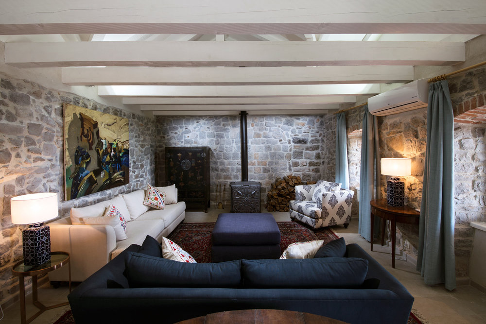 Living room in a villa in Luštica, Montenegro: stone walls, wooden beams, a fireplace, and stylish upholstered furniture.