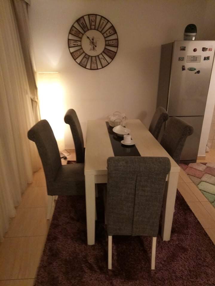 Dining area in the living room of a house in Shushan, Montenegro: a light-colored table, gray chairs, wall clock, and refrigerator.