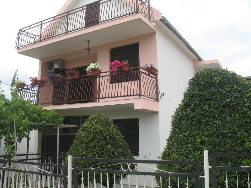 Two-story house with balconies and flowers in Šušanj, Montenegro: building facade with a green garden and wrought-iron fence.