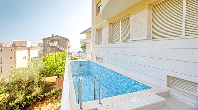 Open-air swimming pool with blue mosaic on the hotel terrace in Bečići, Montenegro, overlooking a residential area and greenery.