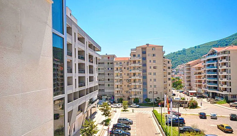 View of urban apartment buildings, a parking lot with cars, and a green hill under a clear blue sky.