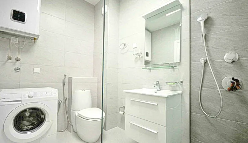 Bathroom with toilet, sink, shower, and washing machine visible in a bright tiled room.