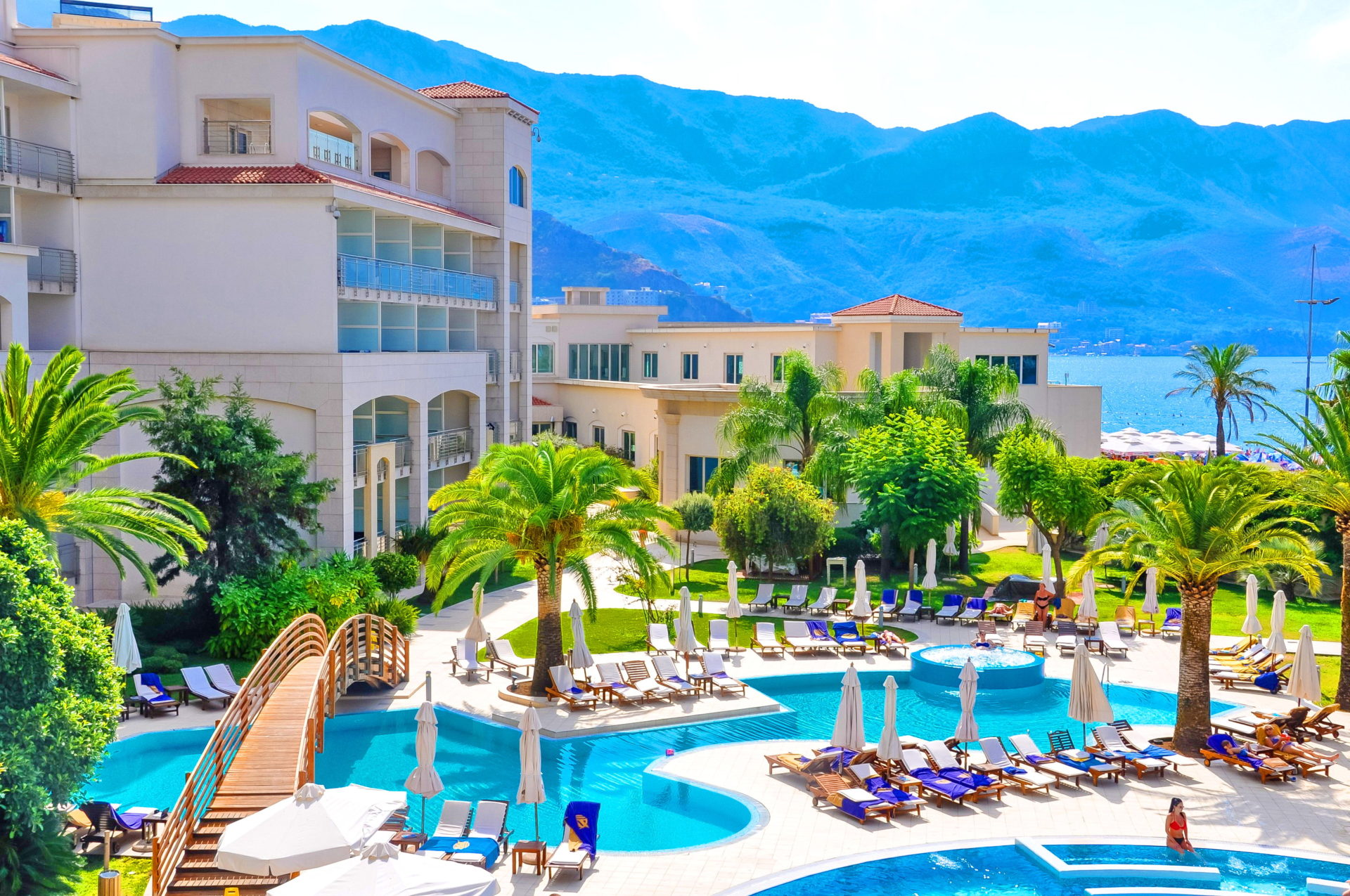 Resort complex with a swimming pool, palm trees, and sea view in Bečići, Montenegro: relaxation area with sun loungers and a bridge.