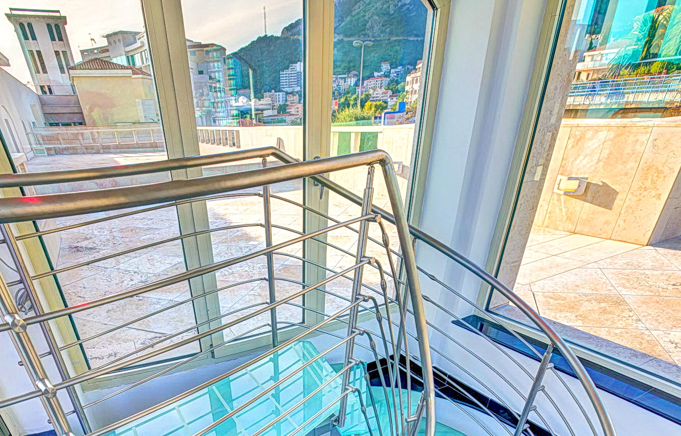 Glass staircase with metal railings and a view of the city of Bečići, Montenegro from the panoramic windows of a modern apartment.