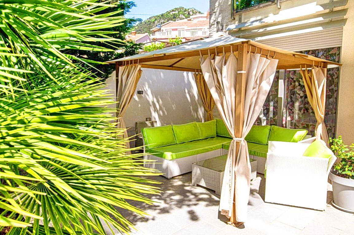 A cozy terrace with a gazebo and wicker furniture at a villa in Budva, Montenegro, surrounded by palm trees and greenery.