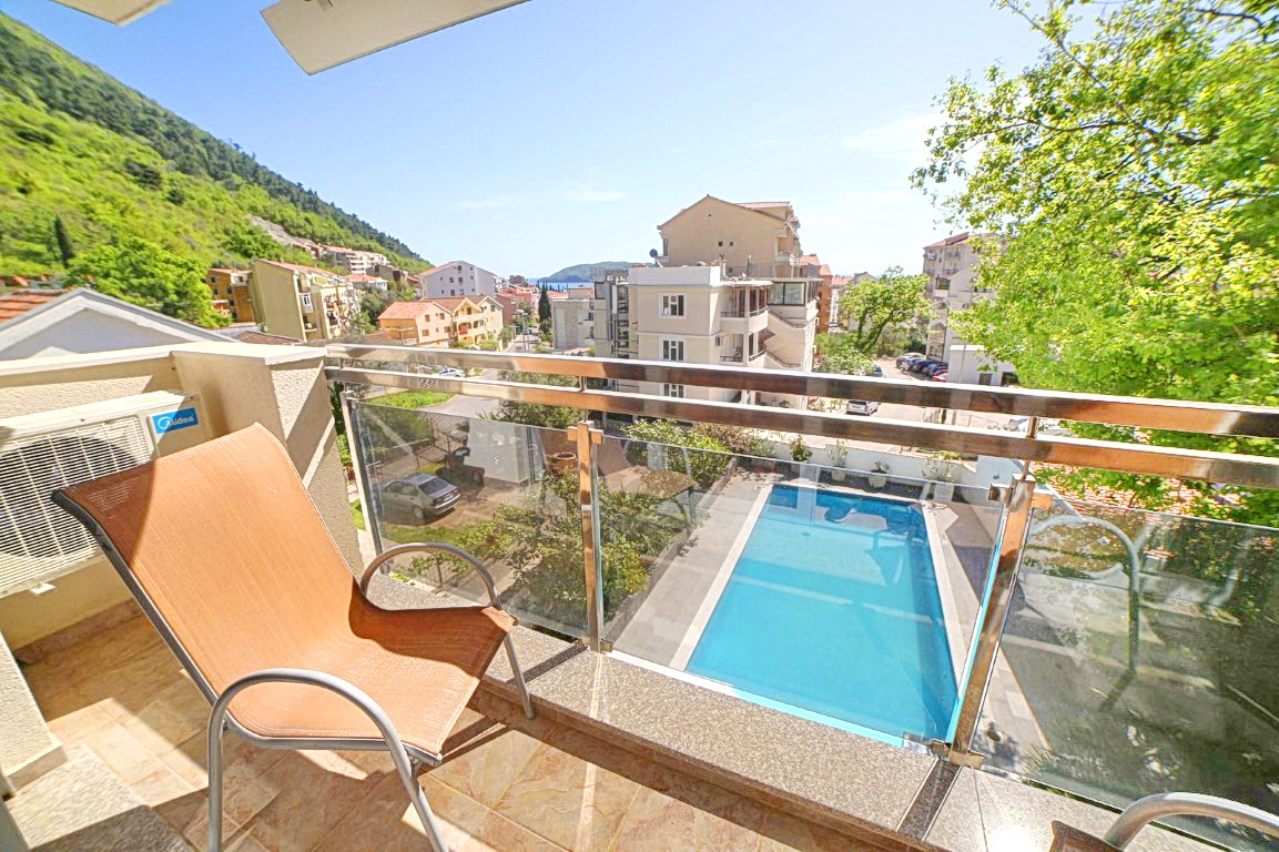 View from the balcony of a villa in Budva, Montenegro: a terrace with a chair, panoramic view of the pool, mountains, and city buildings.