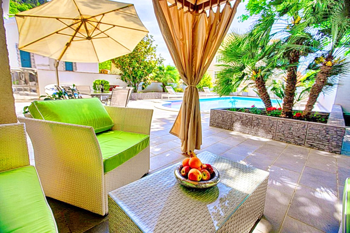 Villa terrace in Budva, Montenegro: wicker furniture, a swimming pool, palm trees, and a sun umbrella in a cozy inner courtyard.