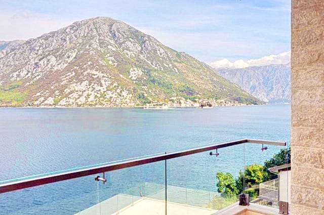Panoramic view of the Bay of Kotor and the mountains from the balcony of a residential complex in Morinj, Montenegro, with a glass railing.