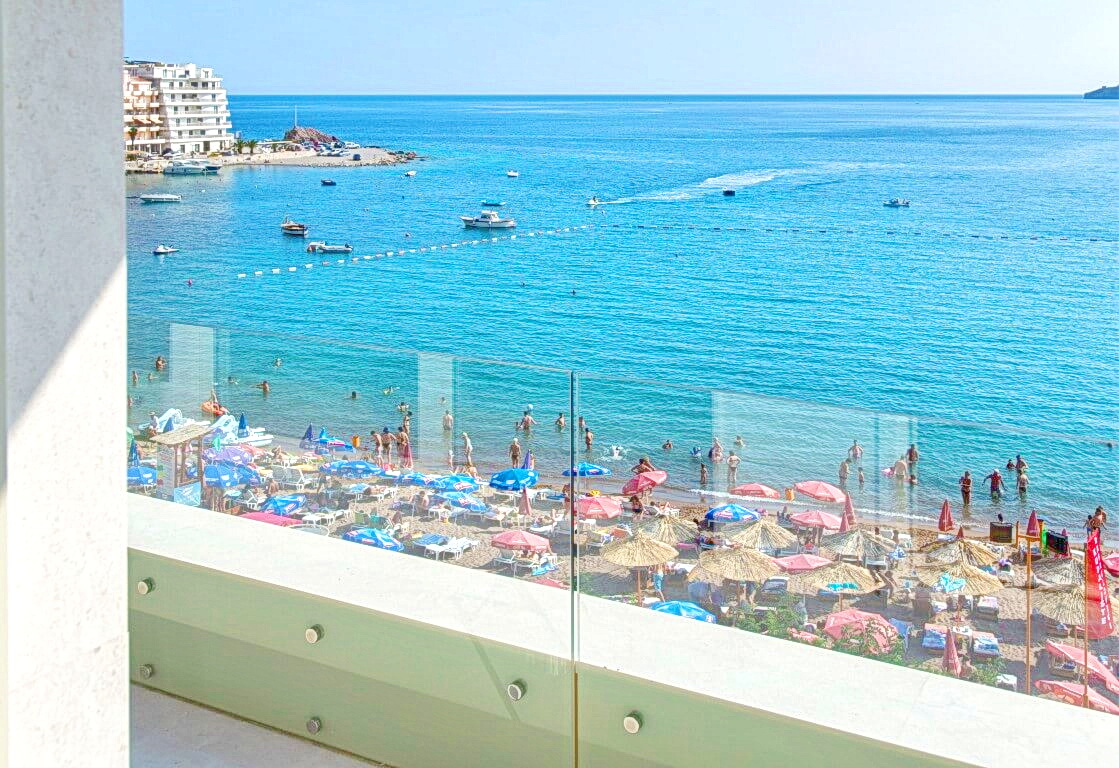 View from the apartment balcony in Rafailovici, Montenegro: a panorama of the beach, the sea, and yachts through a glass railing.