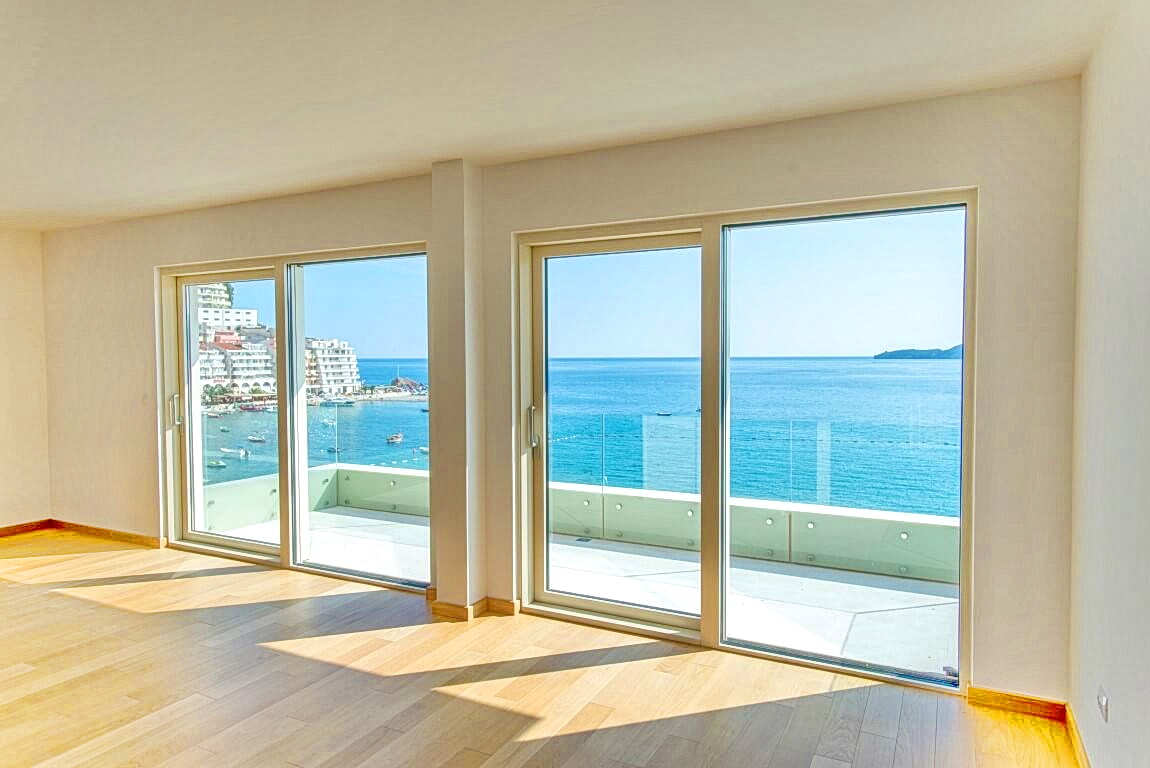 Living room in an apartment in Rafailovići, Montenegro, with panoramic windows, light parquet flooring, and a view of the sea and beach.