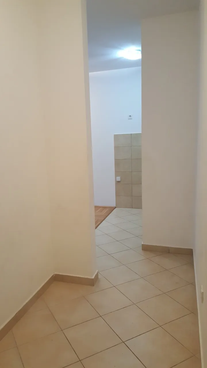 Empty hallway with beige floor tiles and white walls, leading to a room with wooden flooring visible in the background.