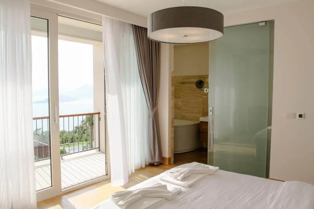 Bedroom with a large bed, white linens, and towels. A glass door leads to a balcony with a sea view.