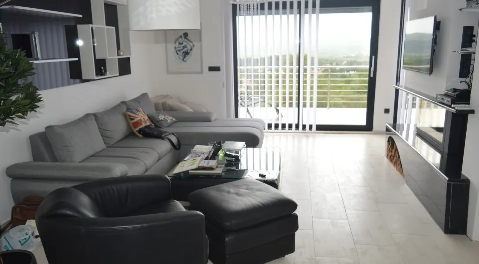 Bright room with a gray sofa, black armchair, glass coffee table, and TV near large windows.