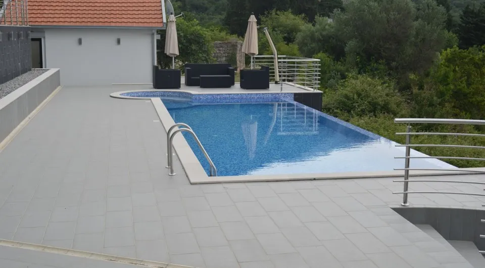 Outdoor swimming pool with blue water surrounded by gray tiles and black patio furniture against greenery.