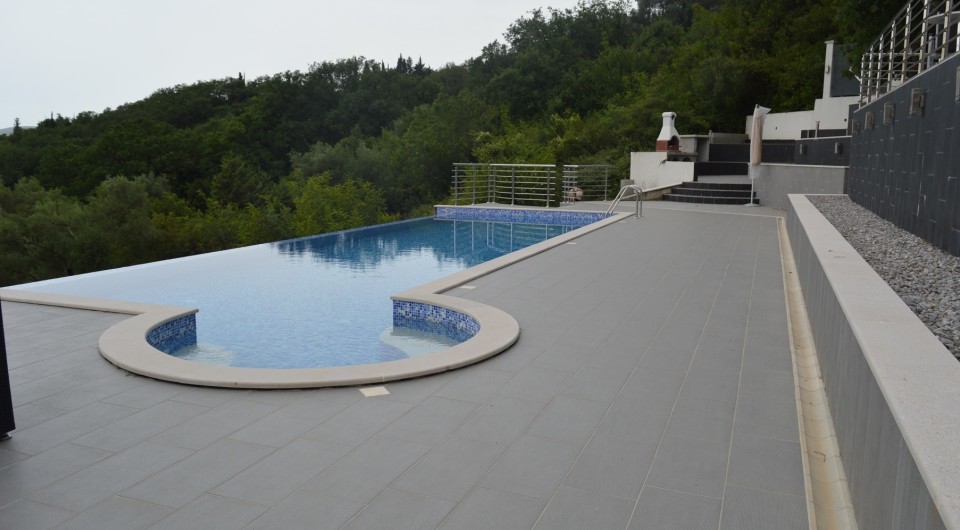Open-air swimming pool with a terrace and barbecue area at a villa in Tivat, Montenegro, against a picturesque forest landscape.