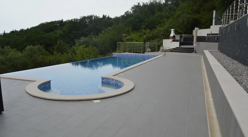 Large swimming pool with blue tile and circular section on gray terrace deck against a backdrop of green hills.