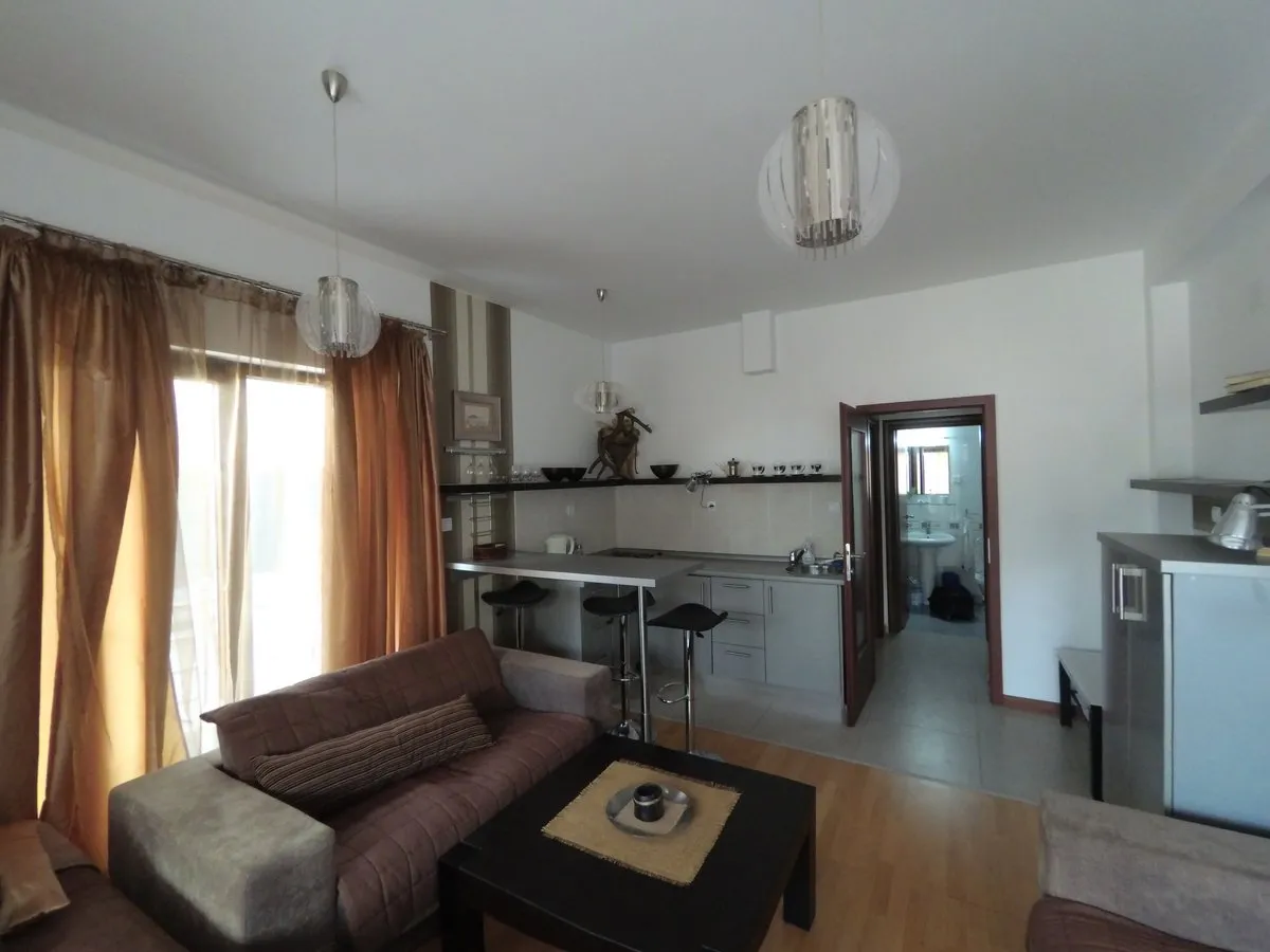 Living room with brown sofa and black table, open kitchen with bar stools and bathroom door visible.