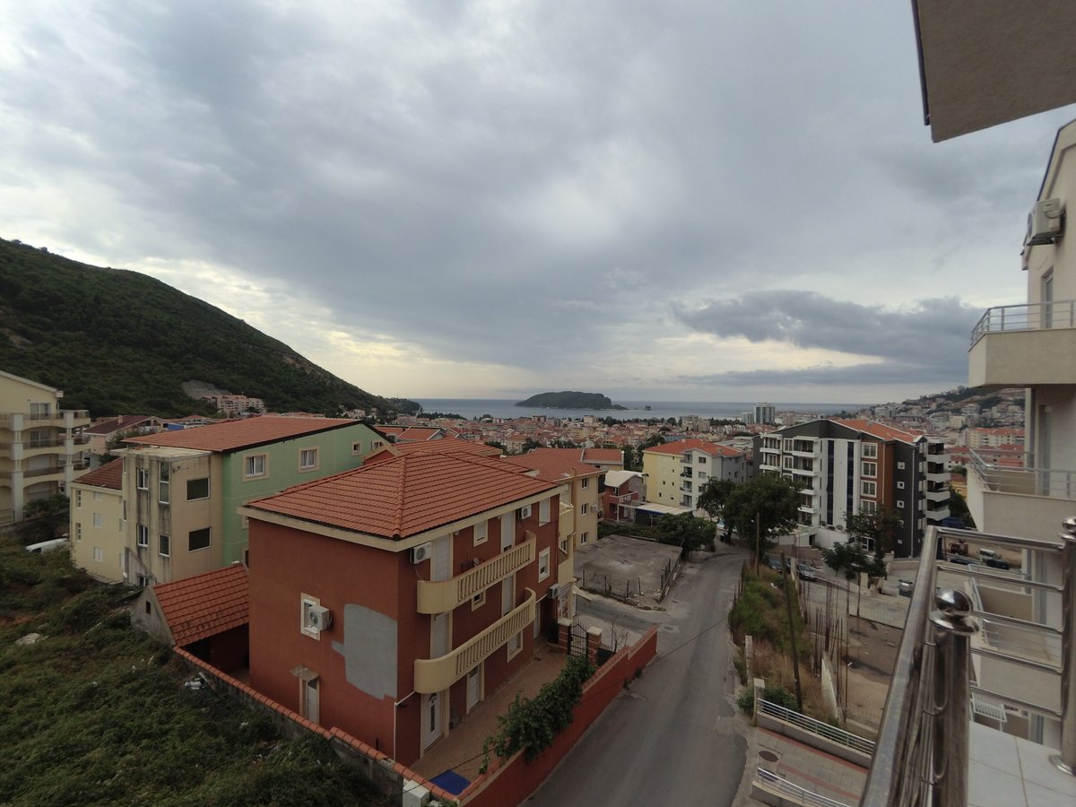 View from the apartment balcony in Budva, Montenegro: a panorama of the city, red tiled roofs, mountains, and the sea bay.