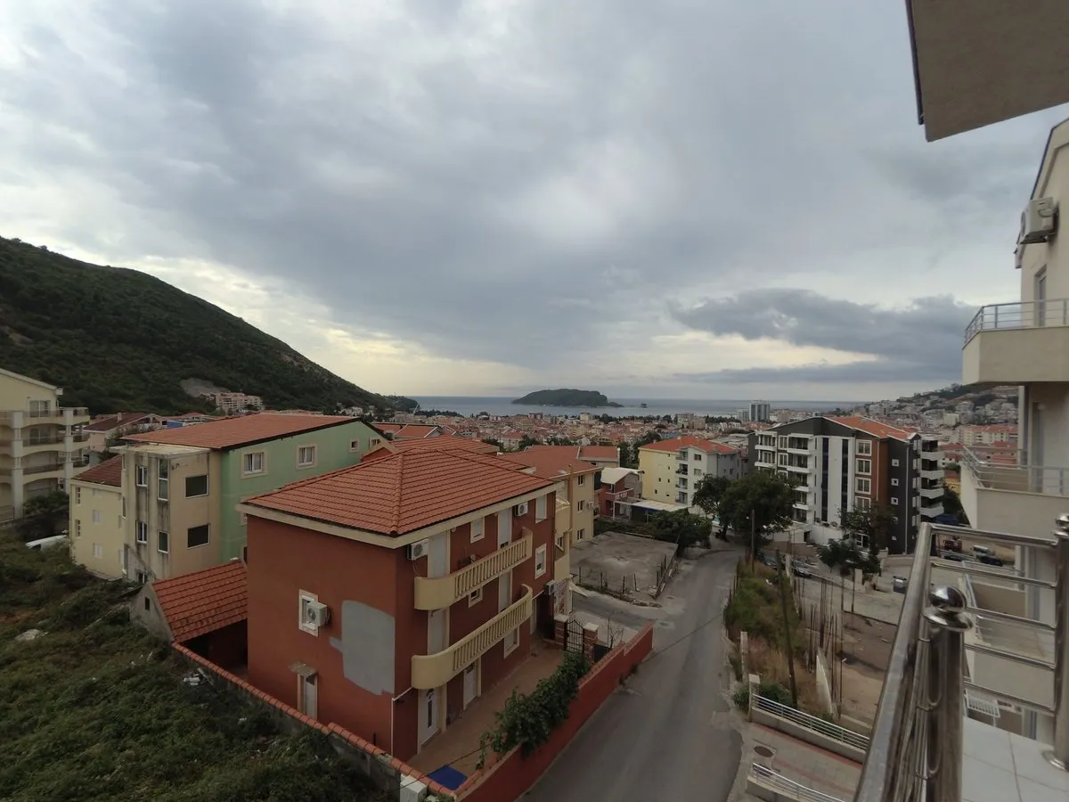 View from a balcony overlooking residential buildings, a street, and the sea under cloudy skies.