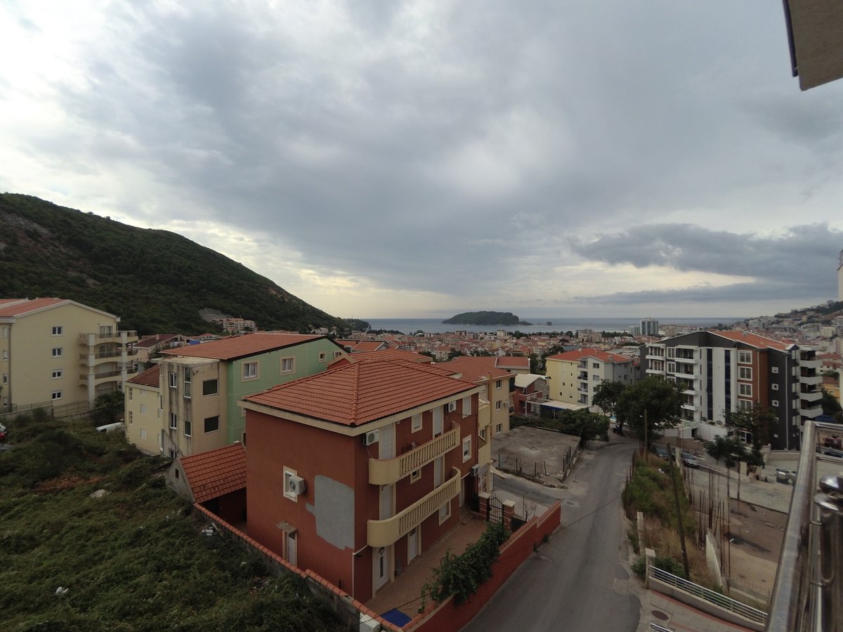 View from the apartment balcony in Budva, Montenegro: a panorama of the city, residential buildings with tiled roofs, and the sea on the horizon.