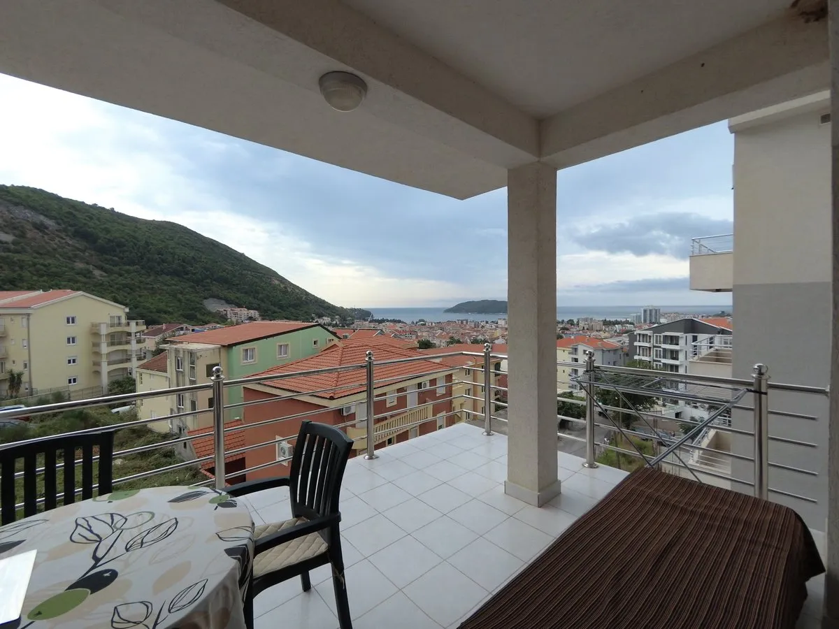 Balcony with city and sea view, tiled floor, metal railing, outdoor table and chair.